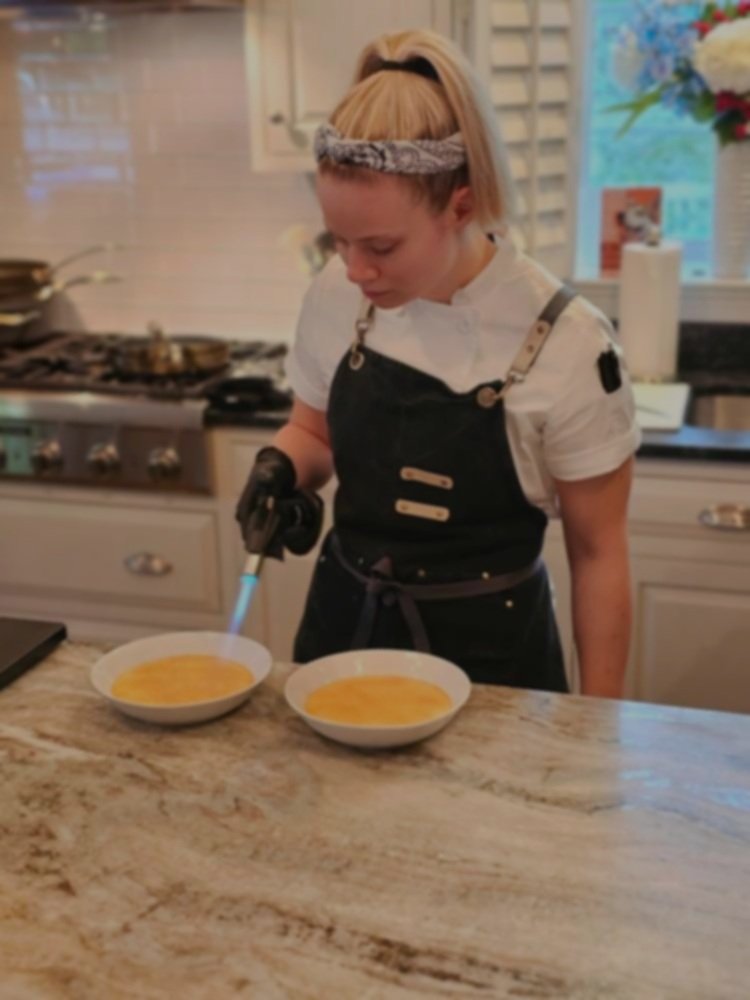 A young girl in a black apron and white shirt uses a kitchen torch to caramelize sugar on two bowls of creme brûlée in a home kitchen.