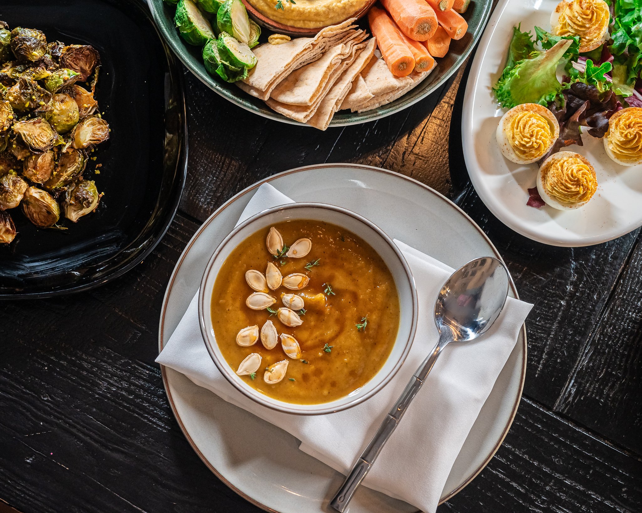 A table set with a bowl of roasted butternut squash soup, roasted Brussels sprouts, a veggie platter with carrots and wraps, and deviled eggs