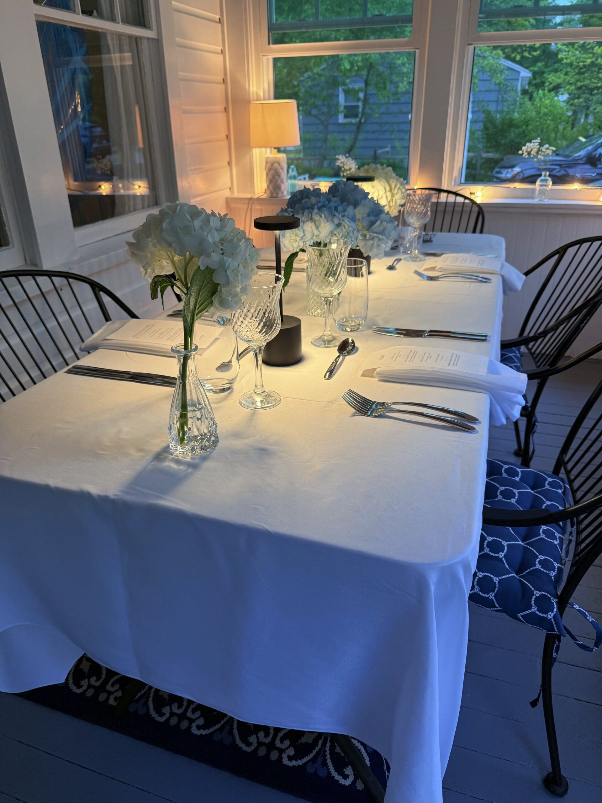 A dinner table set for six with white tablecloth, white floral centerpieces in glass vases, wine glasses, water glasses, silverware, and white napkins, indoors with large windows showing trees outside, warm ambient lighting, and decorative lighting o