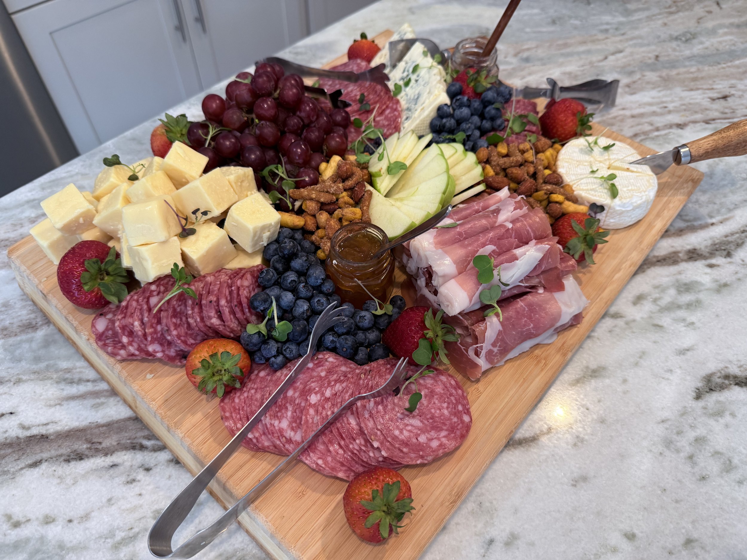 Charcuterie board with cheeses, grapes, strawberries, blueberries, sliced meats, honey, and crackers on a marble countertop.