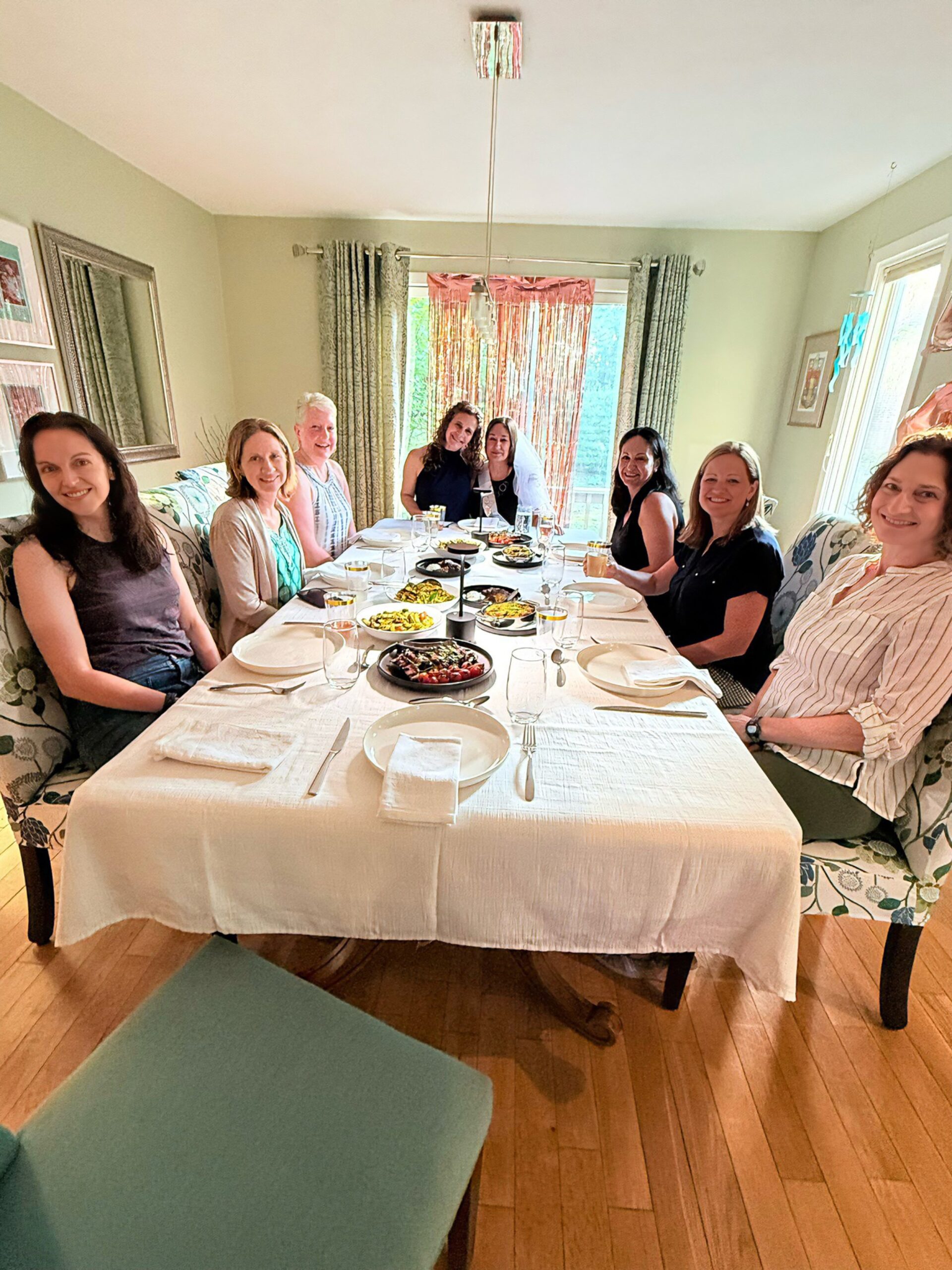 People gathered around a dining table with food and drinks in a well-lit room with curtains and artwork on the walls.