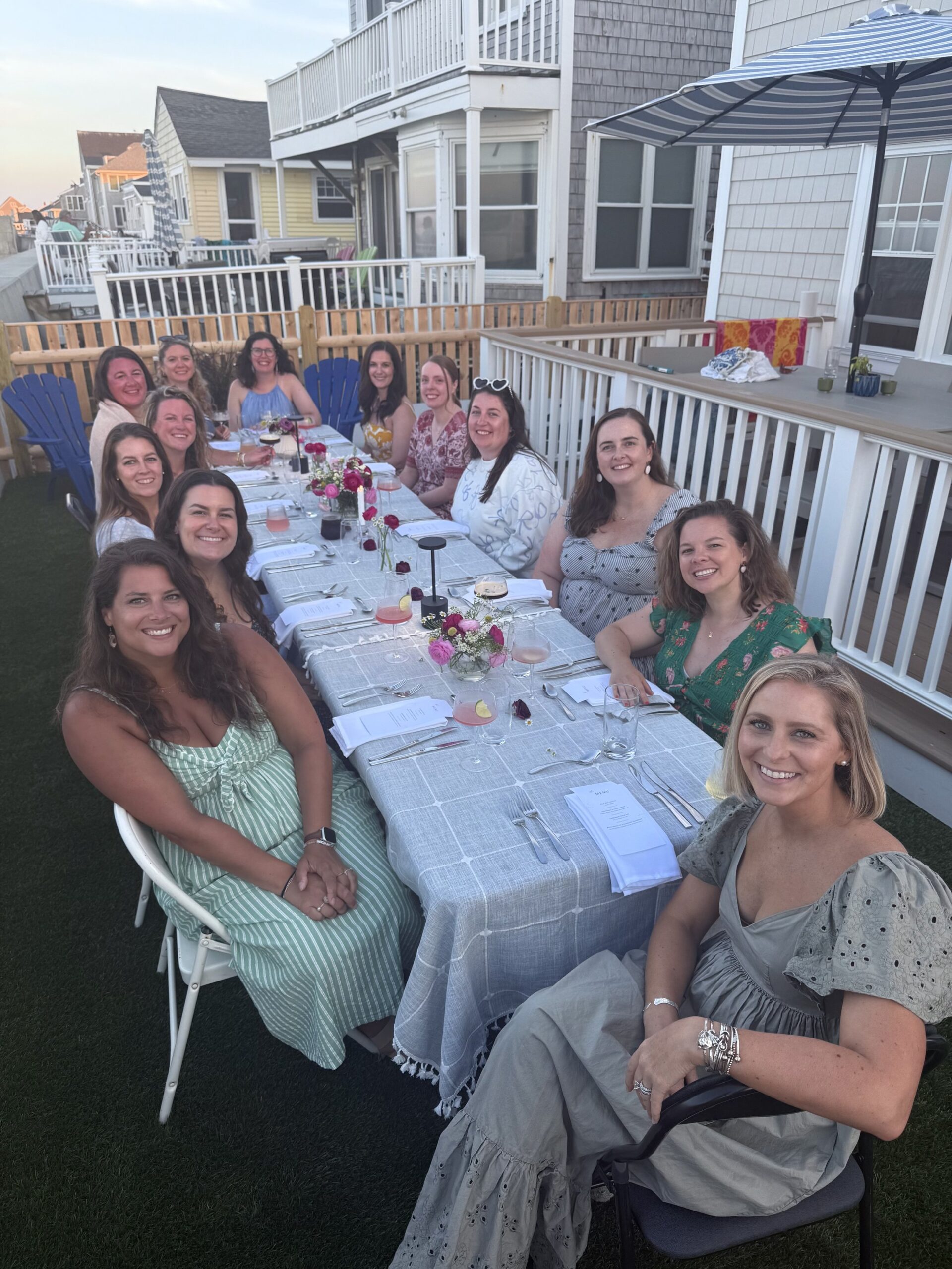 A group of women sitting around a long outdoor dining table on a backyard patio, smiling at the camera during a gathering or celebration, with houses and a fence in the background.