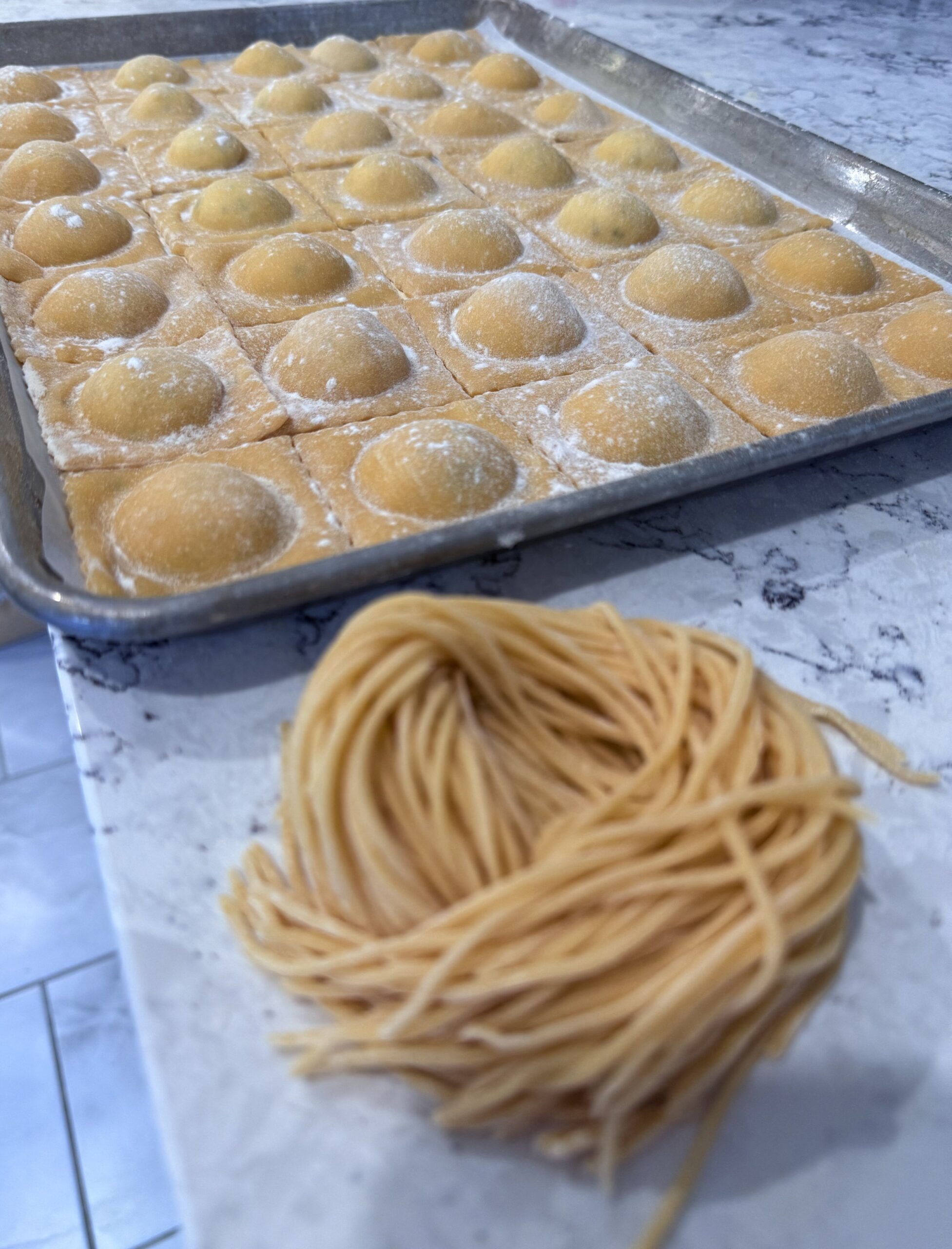 Unbaked ravioli with dome-shaped filling on a baking tray alongside a bundle of uncooked spaghetti on a countertop.